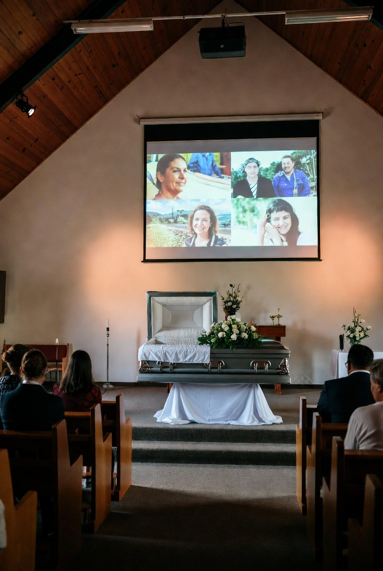 A casket with white flowers at a chapel service while a photo tribute plays on screen